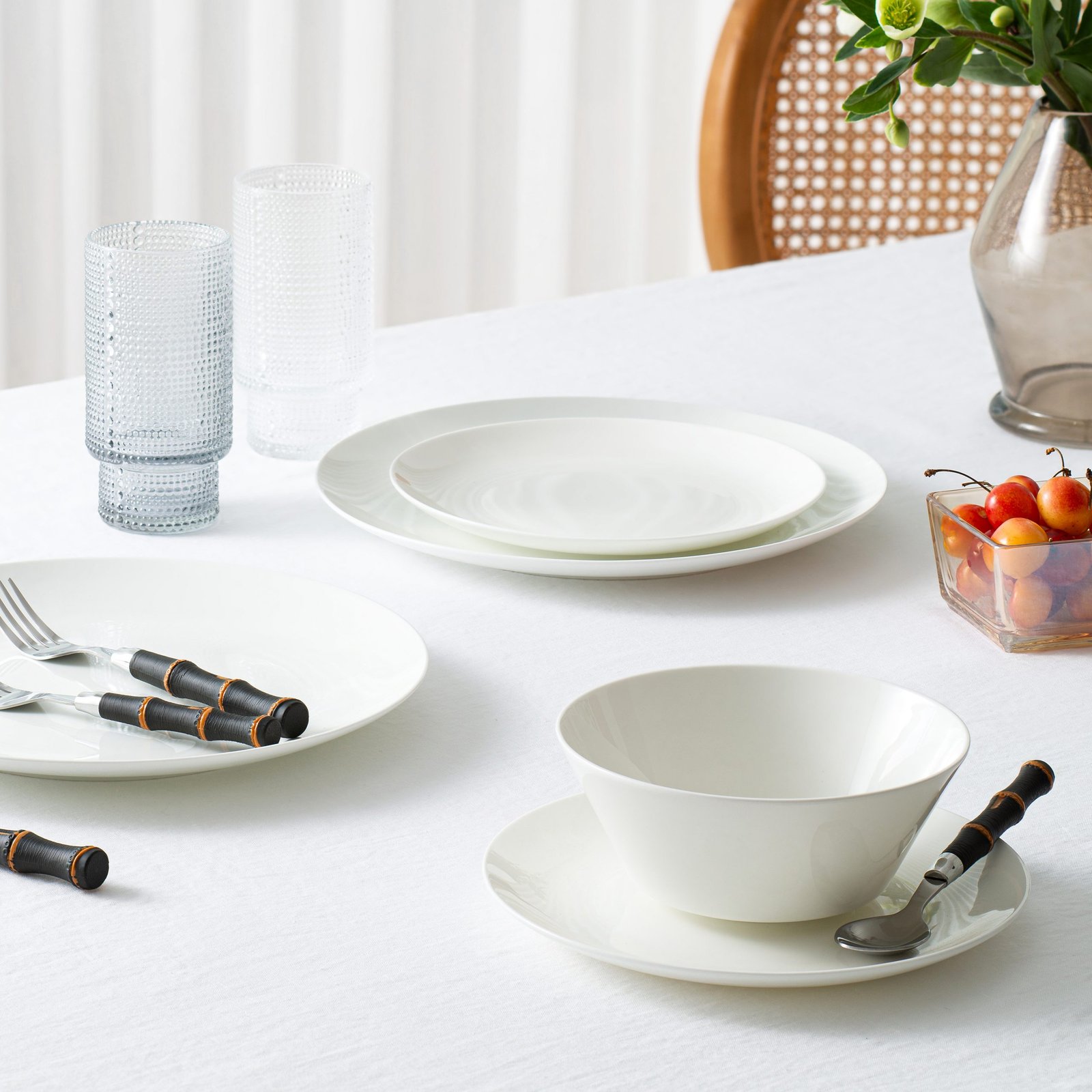 product lifestyle photography of white dinnerware set with wooden handle silverware set white table cloth table with vase basket weave chair and white curtains